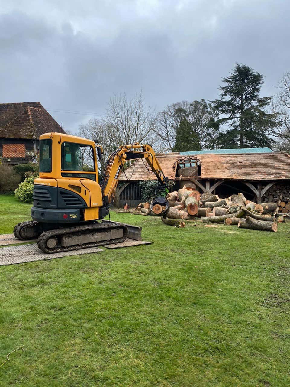 This is a photo of a tree which has grown through the roof of a barn that is being cut down and removed. There is a digger that is removing sections of the tree as well. Bingham Tree Surgeons
